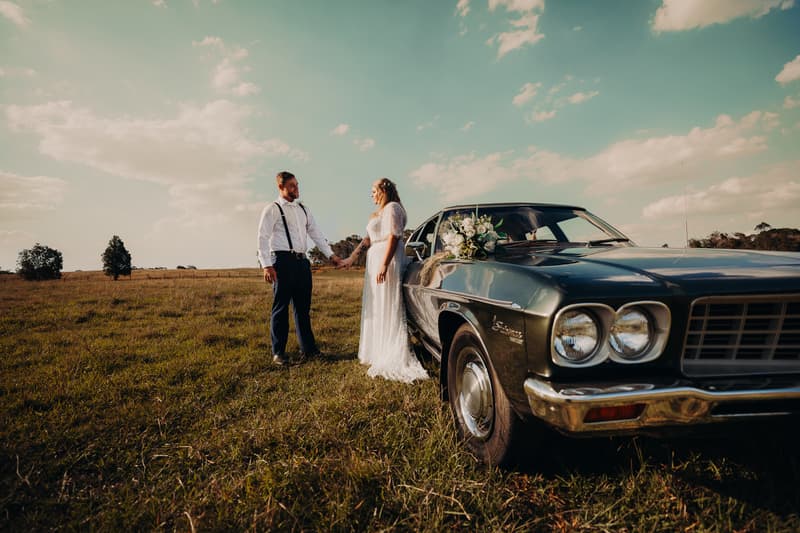 The bride Annie and groom Trent hold hands standing in a grassy field next to a vintage dark green car decorated with a white floral bouquet at Ocean View Estates.