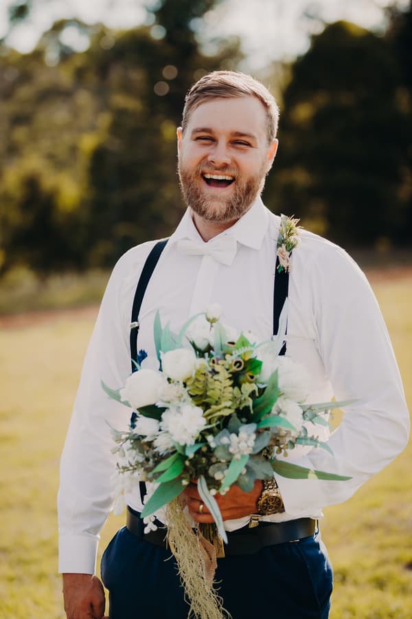The groom Trent stands outdoors at Ocean View Estates holding a bouquet of white and green flowers, wearing a white shirt with black suspenders and a white bow tie, smiling broadly.