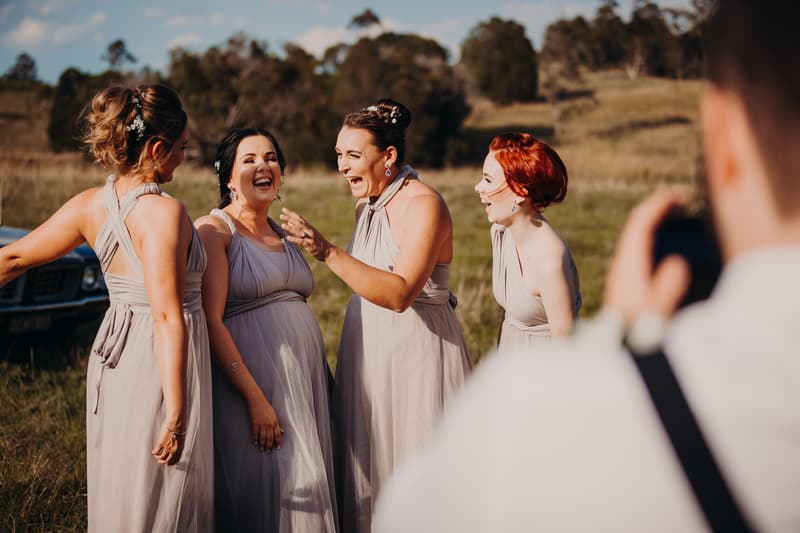 Four bridesmaids in matching light gray dresses laugh together outdoors at Ocean View Estates, with a man in the foreground taking their photo.