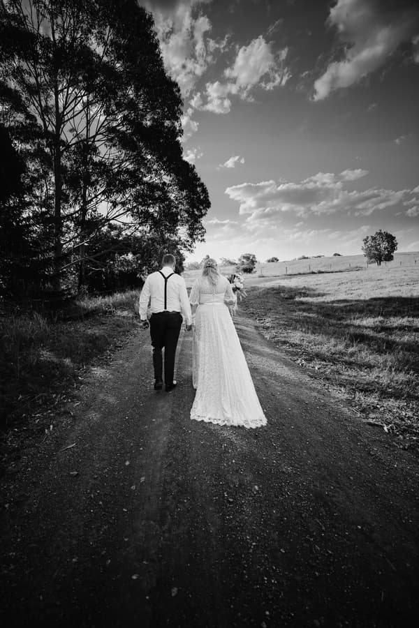 The bride Annie and groom Trent walk hand in hand down a dirt path at Ocean View Estates, with trees on the left and open fields on the right under a partly cloudy sky.