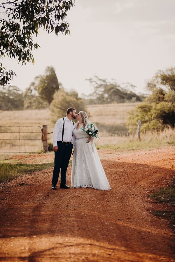 The bride Annie and groom Trent kiss while standing on a dirt path at Ocean View Estates. Annie wears a white lace wedding dress and holds a bouquet, while Trent wears dark pants, a white shirt, and suspenders. The background shows a rural landscape with trees and a metal gate.