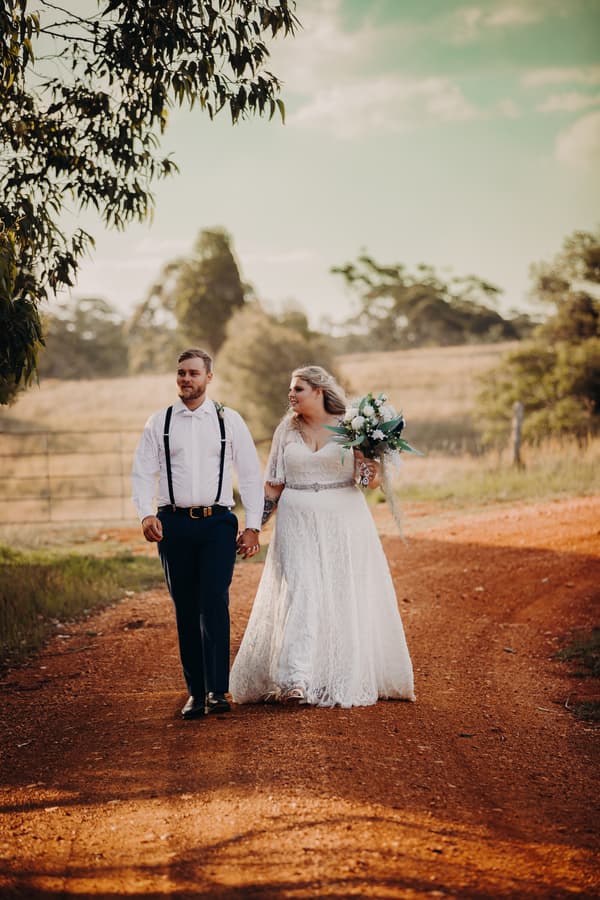The bride Annie in a white lace wedding gown holding a bouquet and the groom Trent in a white shirt with suspenders and dark pants walk hand in hand along a dirt path at Ocean View Estates.