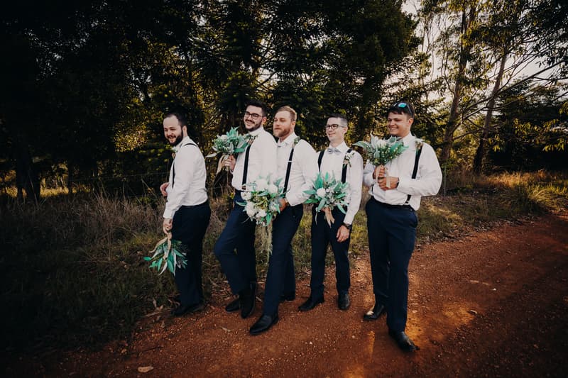 Five groomsmen stand on a dirt path holding bouquets in front of trees at Ocean View Estates.
