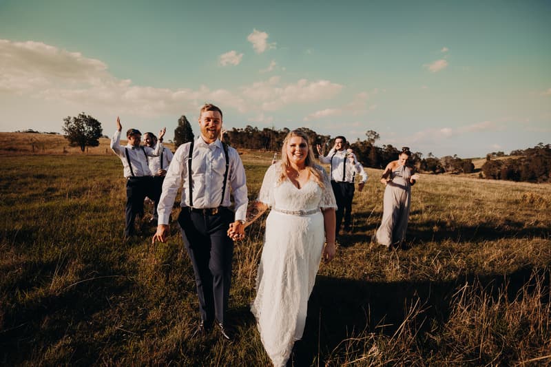 Bride Annie and groom Trent walk hand in hand across a grassy field at Ocean View Estates, followed by their wedding party dressed in coordinated attire.