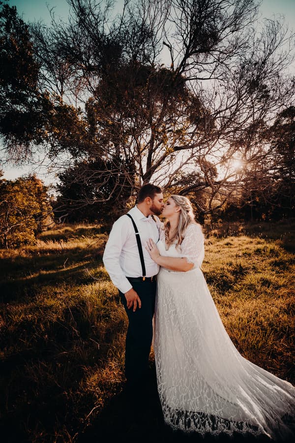 The bride Annie and groom Trent share a kiss outdoors at Ocean View Estates during their couple portraits session, standing in a grassy area with trees and the sun low in the sky behind them.