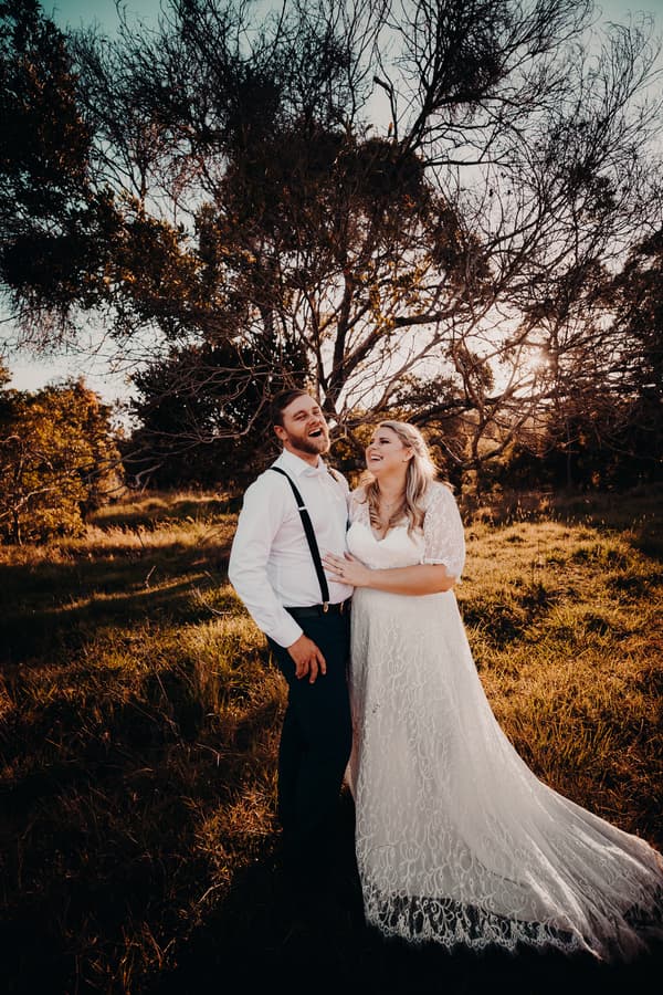 Annie in a white lace wedding dress and Trent in a white shirt with black suspenders stand together outdoors at Ocean View Estates, surrounded by grass and trees with sunlight filtering through.