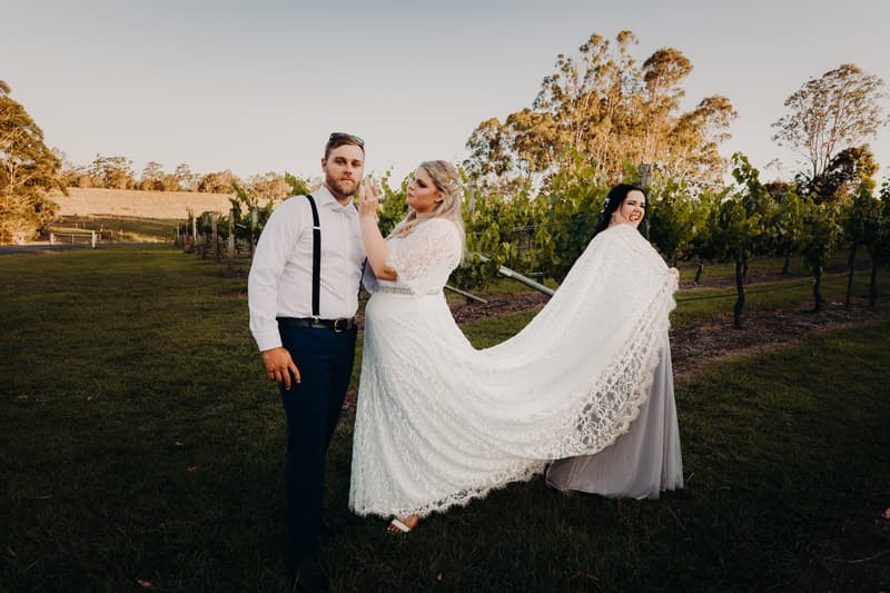 The bride Annie and groom Trent stand outdoors at Ocean View Estates with a bridesmaid holding the bride's lace train behind her among grapevines.