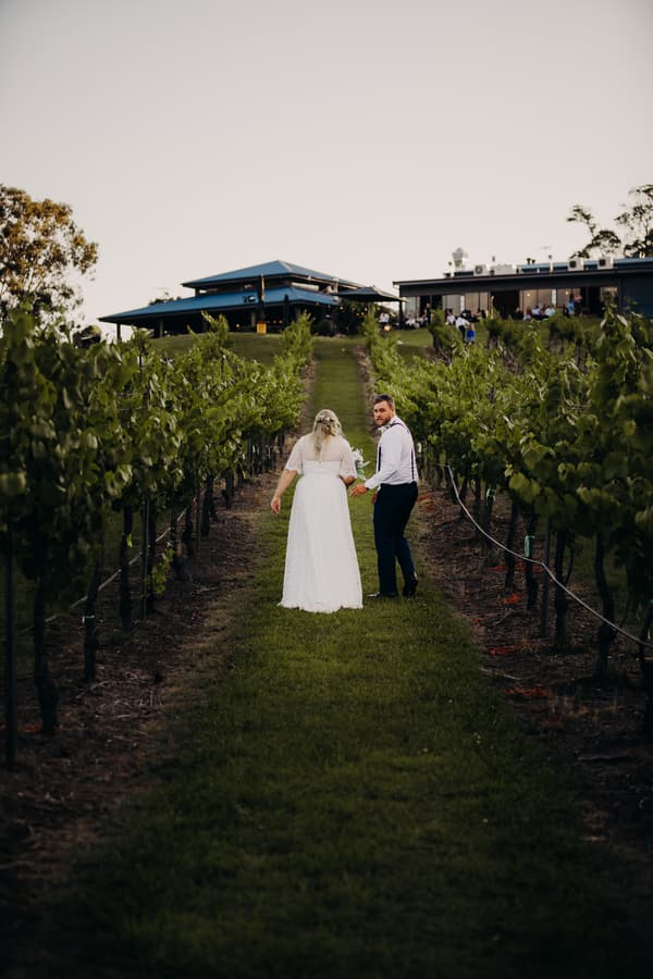 The bride Annie and groom Trent walk hand in hand through a vineyard at Ocean View Estates, with the estate building visible in the background.