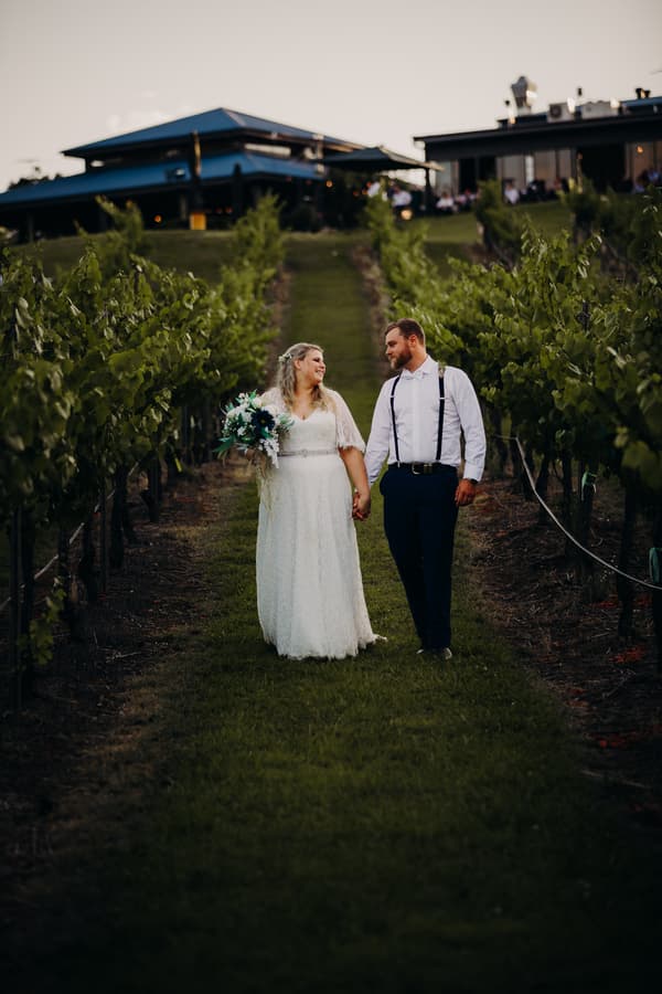 Annie the bride and Trent the groom walk hand in hand down a grassy path between rows of grapevines at Ocean View Estates, with estate buildings visible in the background.
