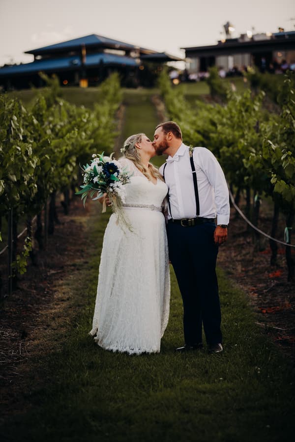 The bride Annie and groom Trent kiss while standing in a vineyard at Ocean View Estates. Annie wears a white lace wedding dress and holds a bouquet, while Trent wears a white shirt with suspenders and dark pants.