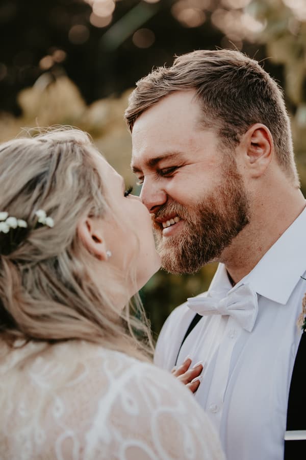Annie the bride and Trent the groom share a close moment during their couple portraits at Ocean View Estates. Annie wears a lace wedding dress with floral hair accessories, and Trent wears a white shirt with a bow tie and suspenders.