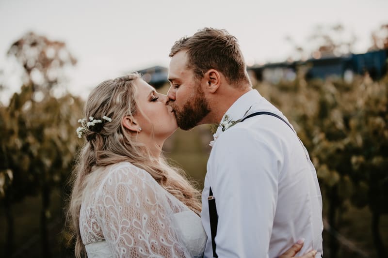 The bride Annie and groom Trent share a kiss outdoors, surrounded by greenery and vineyard rows. May wedding sunset Brisbane by She Said Yes.