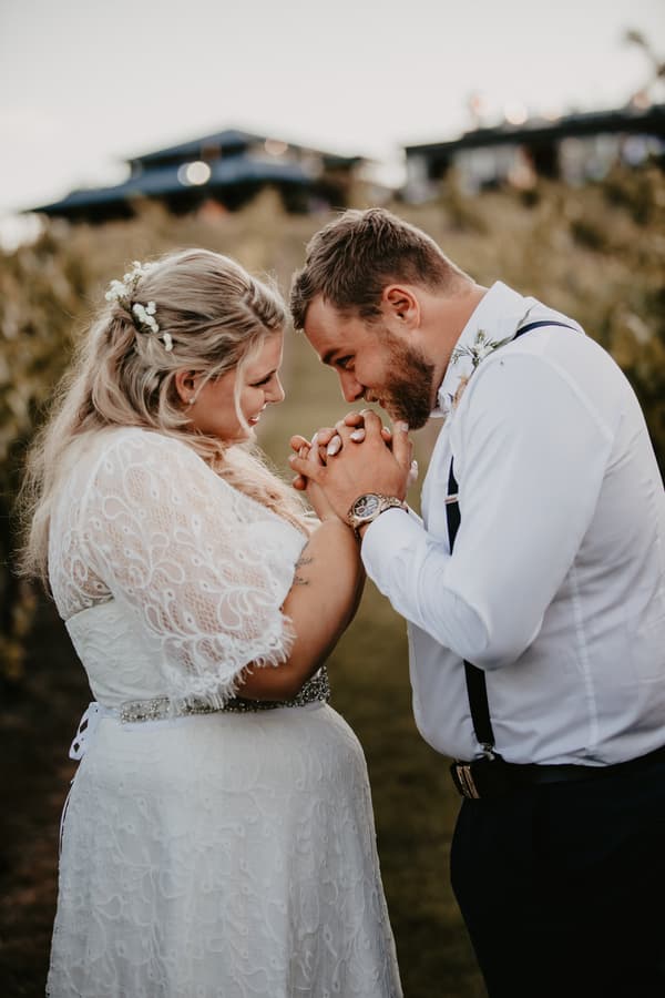 The bride Annie and groom Trent stand facing each other holding hands closely at Ocean View Estates, with greenery and buildings blurred in the background.