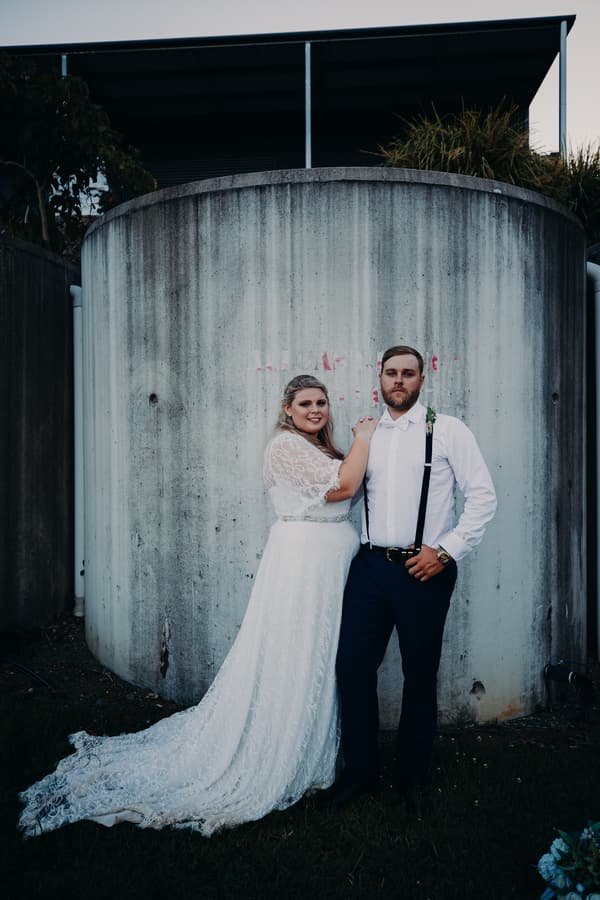 The bride Annie in a white lace wedding gown and the groom Trent in a white shirt with suspenders pose together in front of a large cylindrical concrete structure at Ocean View Estates.