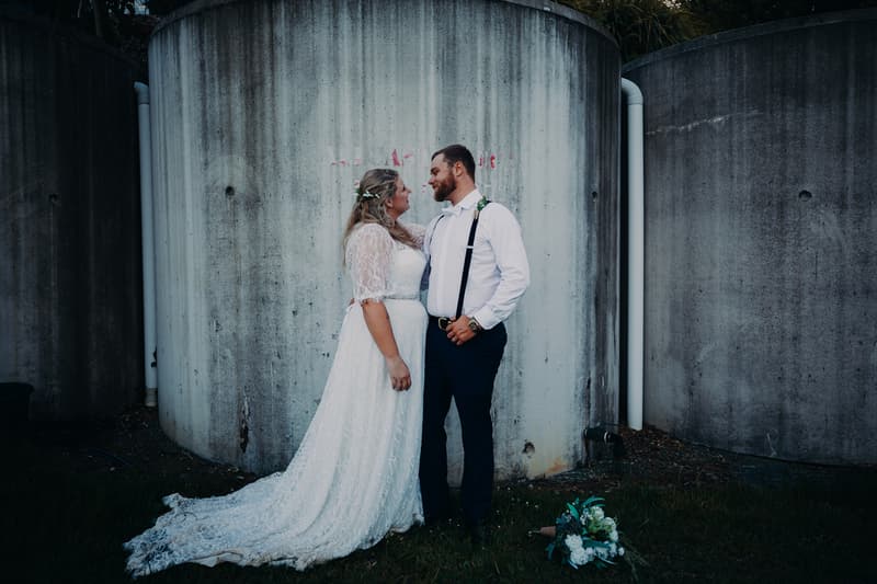 The bride Annie in a lace wedding dress and the groom Trent in a white shirt with suspenders stand facing each other near large concrete cylindrical structures at Ocean View Estates. A bridal bouquet lies on the grass nearby.
