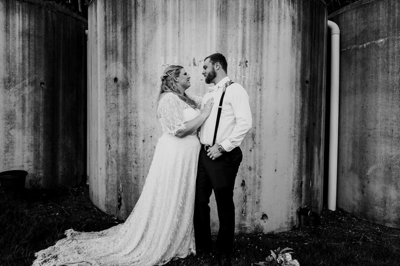 The bride Annie and groom Trent stand facing each other in front of large cylindrical concrete structures at Ocean View Estates, with Annie wearing a lace wedding dress and Trent in a white shirt with suspenders.