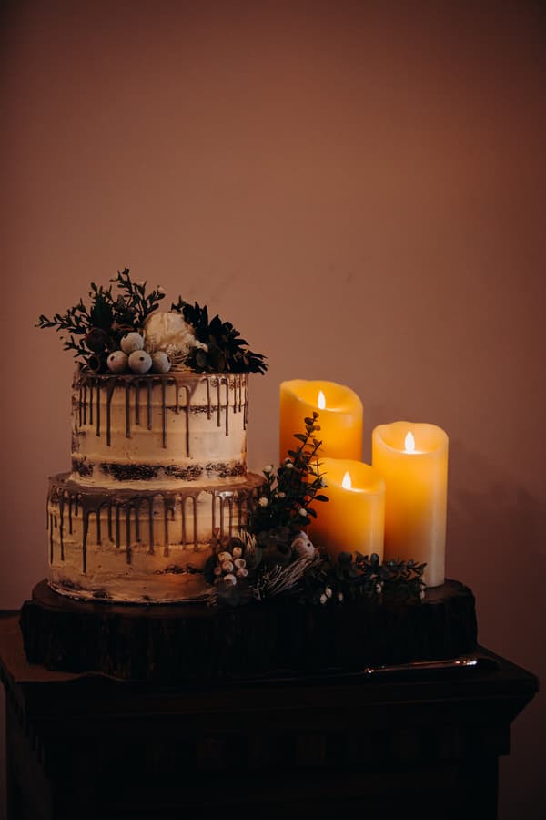 Two-tier wedding cake decorated with greenery and berries next to three lit candles on a wooden slab at Ocean View Estates.