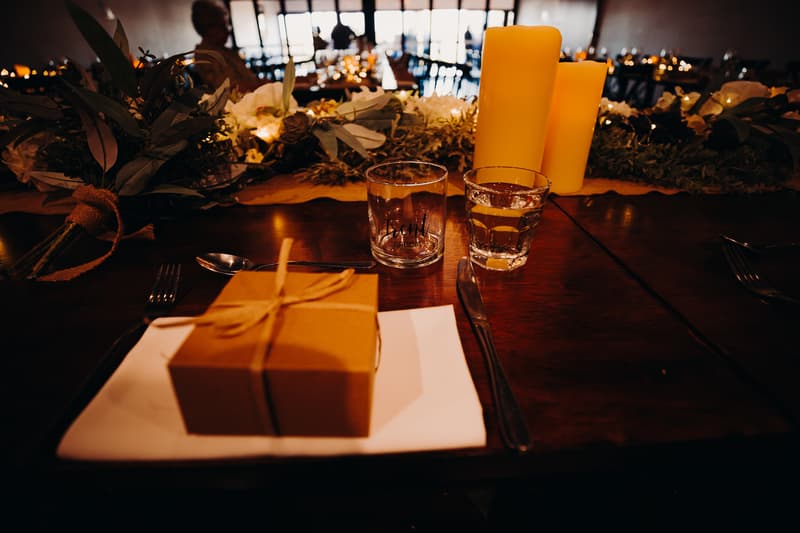 A reception table at Ocean View Estates set with a wrapped gift box on a napkin, cutlery, two glasses including one labeled 'GROOM', and decorative candles and floral arrangements in the background.