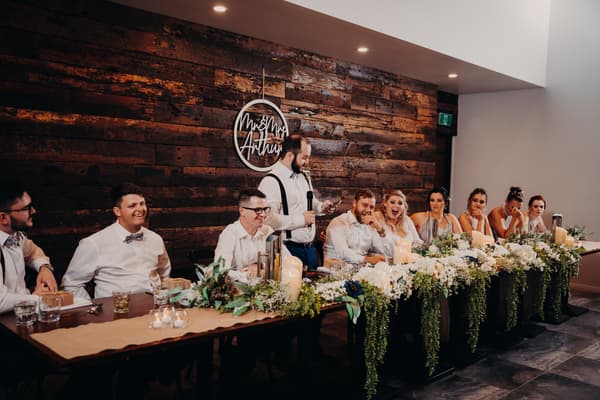 The bridal party sits at a long table decorated with white flowers and greenery at Ocean View Estates. A man in suspenders stands holding a microphone, appearing to give a speech or toast. The group includes bridesmaids in matching dresses and groomsmen in white shirts and bow ties.