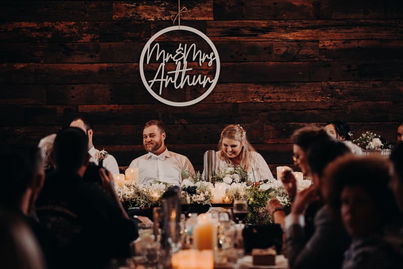 Bride Annie and groom Trent sit at the head table during the reception at Ocean View Estates, surrounded by floral arrangements and candles, with guests seated at tables in the foreground.