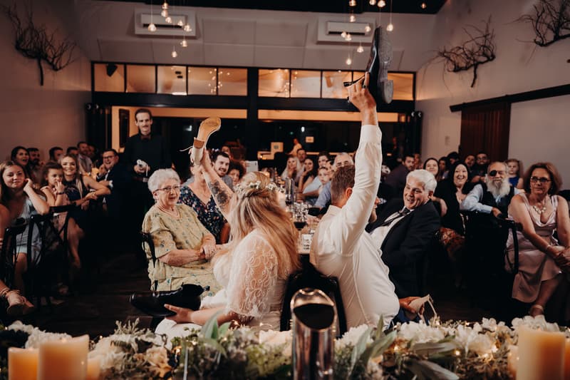 Bride Annie and groom Trent participate in the shoe game at their wedding reception at Ocean View Estates, seated back-to-back holding up each other's shoes while guests watch and laugh.