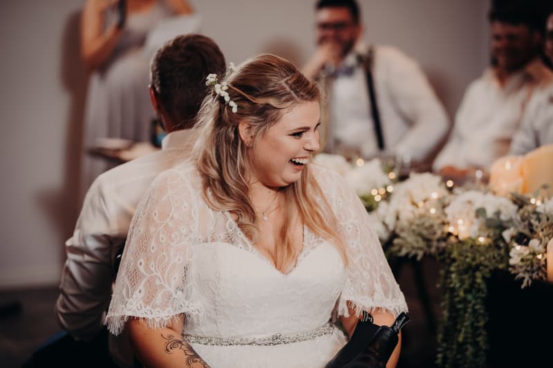 The bride Annie laughs while seated back-to-back with the groom Trent at Ocean View Estates reception stage, with guests and floral decorations visible in the background.