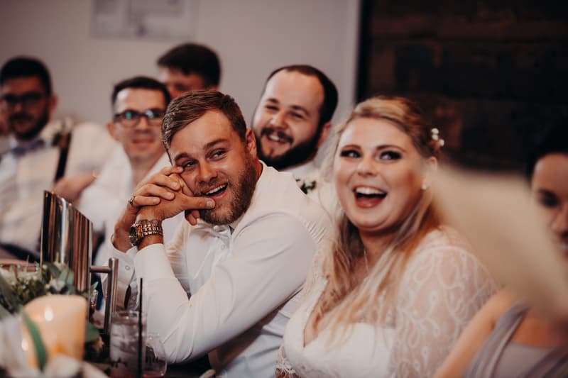The groom Trent and the bride Annie sit at a reception table at Ocean View Estates, surrounded by guests, smiling and enjoying the moment.
