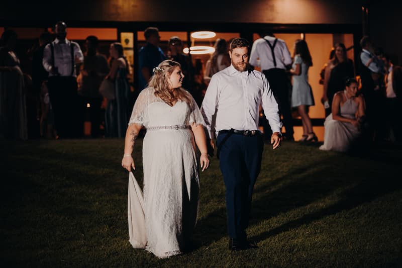 The bride Annie in a white lace gown and the groom Trent in a white shirt and dark pants walk hand in hand on the grass at Ocean View Estates during the reception, with guests standing and sitting in the background near a building entrance.