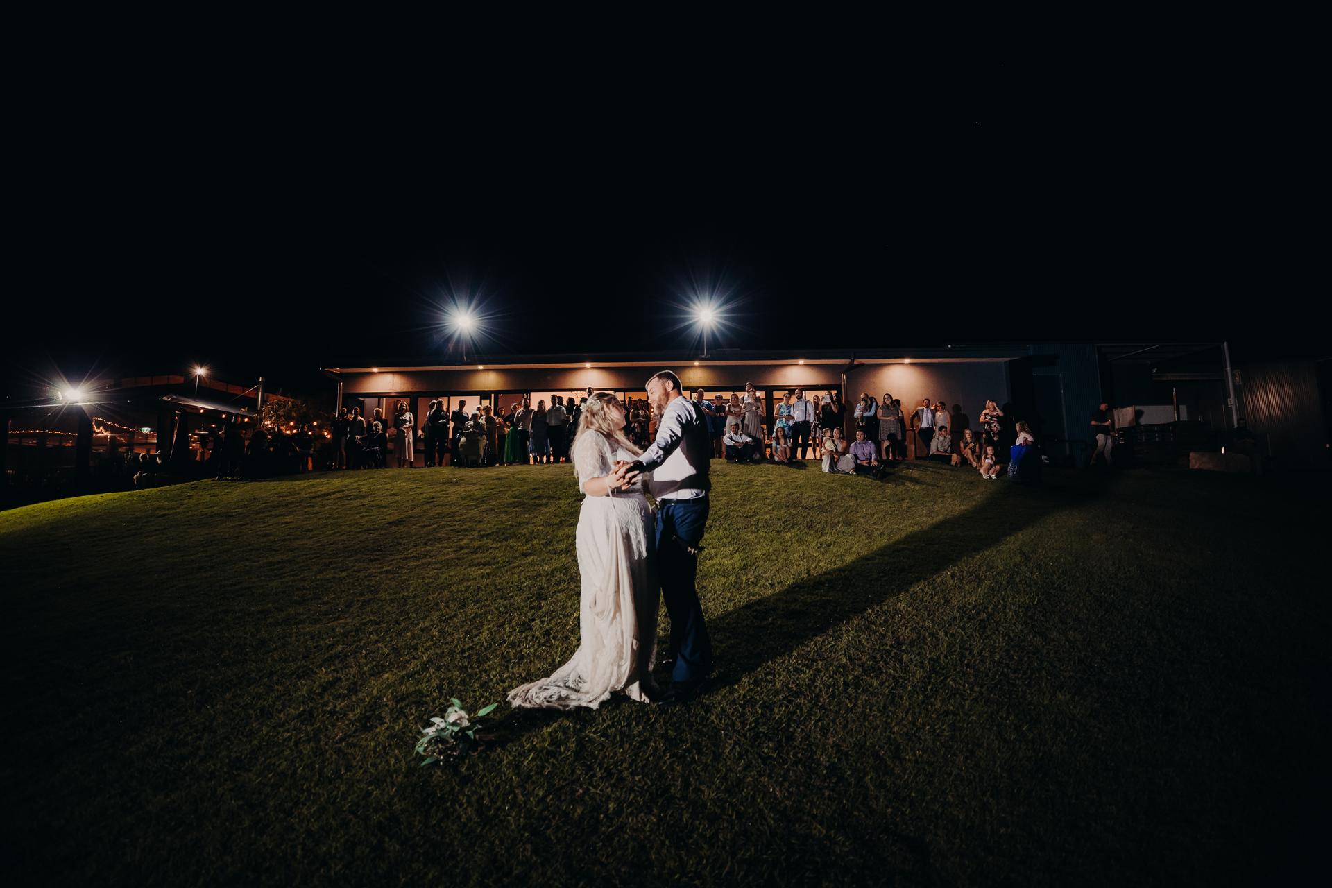 The bride Annie and groom Trent share a dance on a grassy hill at night while guests watch from the lit building behind them at Ocean View Estates.