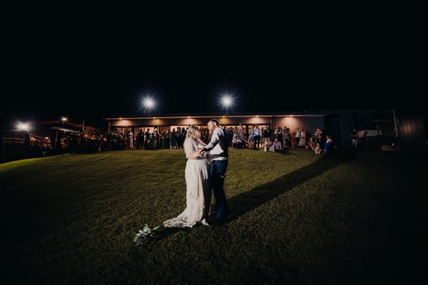 The bride Annie and groom Trent share a dance on a grassy hill at night while guests watch from the lit building behind them at Ocean View Estates.