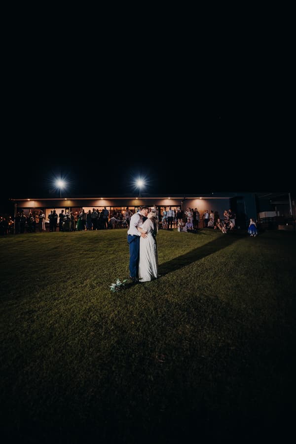 Bride Annie and groom Trent share a kiss on the grass at Ocean View Estates during the reception, with guests watching from a lit building in the background at night.