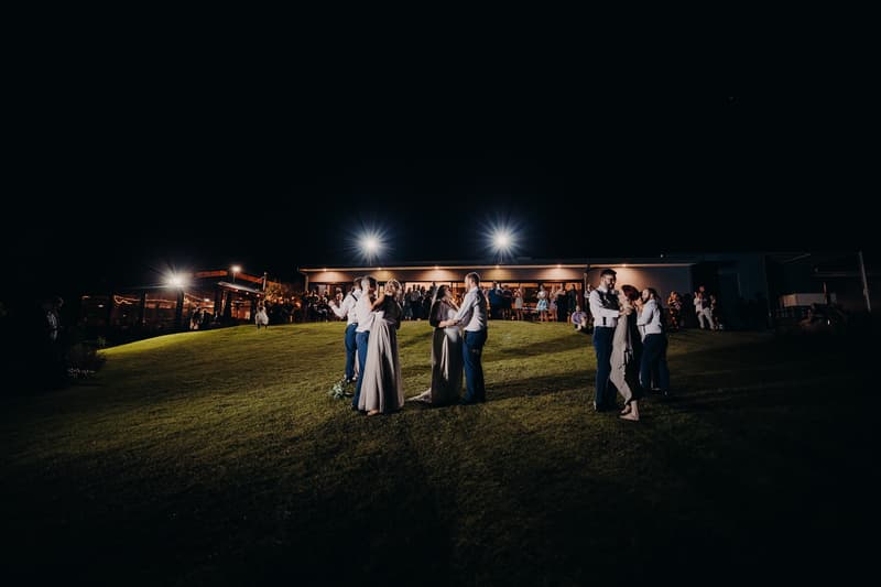 At Ocean View Estates, three couples including the bride and groom dance on a grassy slope outside the reception building at night, with guests visible near the lit structure in the background.