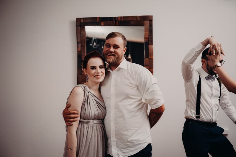 The bride and groom pose together in front of a mirror at Ocean View Estates during the reception stage, with another guest dancing nearby.