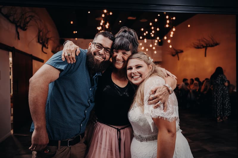 The bride Annie, the groom Trent, and an older woman embrace and smile together at the reception stage of Ocean View Estates.