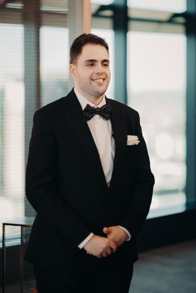 The groom Antony stands indoors at Brisbane registry ceremony room wearing a black suit with a white shirt and a black polka dot bow tie, smiling and looking slightly to the side.