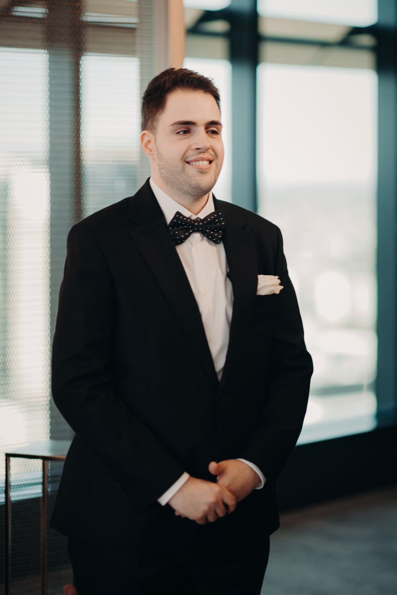 The groom Antony stands indoors at Brisbane registry ceremony room wearing a black suit with a white shirt and a black polka dot bow tie, smiling and looking slightly to the side.