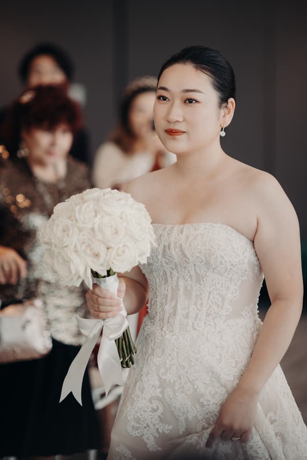 The bride Aria holds a bouquet of white roses at Brisbane registry ceremony room with guests blurred in the background.