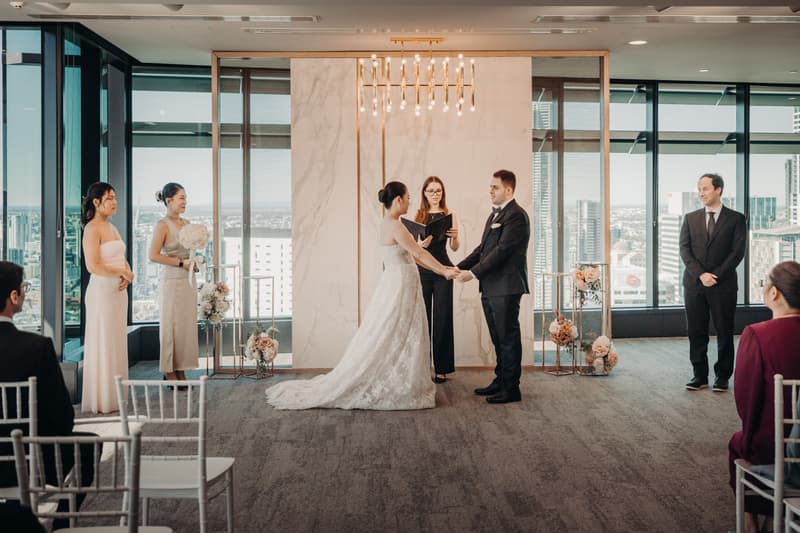 Bride Aria and groom Antony hold hands facing each other during their wedding ceremony at Brisbane registry ceremony room, with the officiant reading vows between them, two bridesmaids standing to the left, a groomsman to the right, and seated guests partially visible.