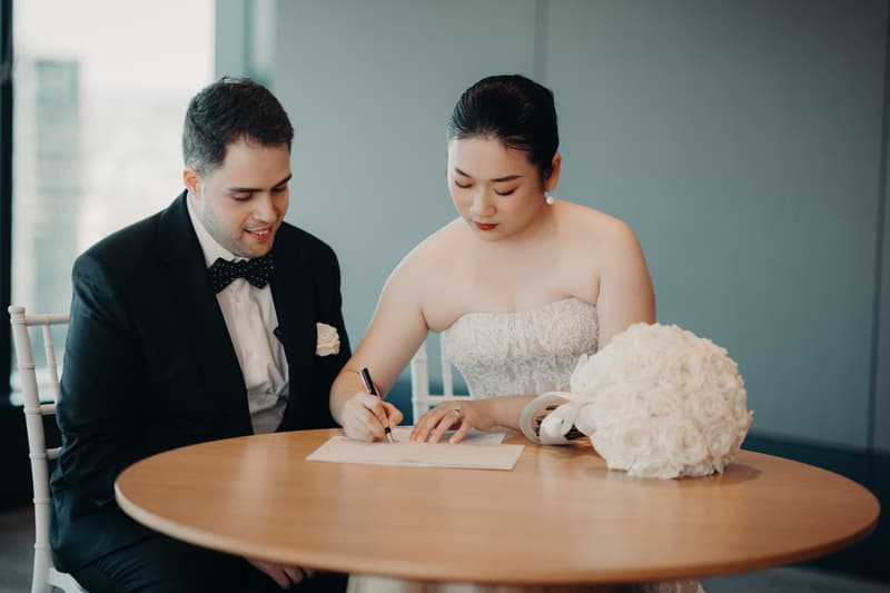 The bride Aria signs a document at a round wooden table while the groom Antony looks on at Brisbane registry ceremony room. A white bridal bouquet rests on the table.