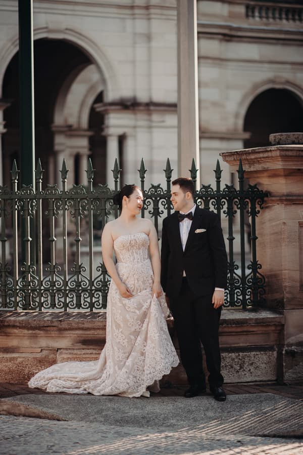 The bride Aria in a strapless lace wedding gown and the groom Antony in a black tuxedo with a bow tie stand side by side in front of an ornate iron fence at the Queensland Parliament building.