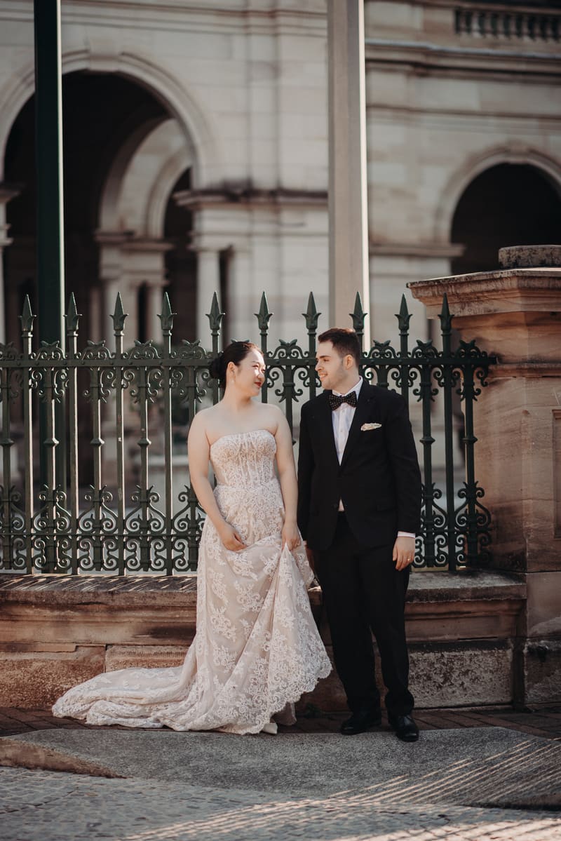 The bride Aria in a strapless lace wedding gown and the groom Antony in a black tuxedo with a bow tie stand side by side in front of an ornate iron fence at the. May wedding sunset Brisbane by She Said Yes.