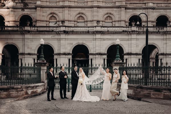 Aria and Antony pose for couple portraits with their wedding party outside the Queensland Parliament building. Aria wears a lace wedding gown and holds a bouquet, Antony is in a black tuxedo. Three bridesmaids in light-colored dresses and two groomsmen in black suits stand beside them.