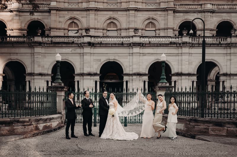 Aria and Antony pose for couple portraits with their wedding party outside the Queensland Parliament building. Aria wears a lace wedding gown and holds a bouquet, Antony is in a black tuxedo. Three bridesmaids in light-colored dresses and two groomsmen in black suits stand beside them.