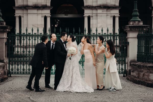 Aria and Antony stand in front of a green wrought iron fence at Queensland Parliament building, surrounded by three bridesmaids in light-colored dresses and two groomsmen in black suits, posing playfully with the bride and groom.