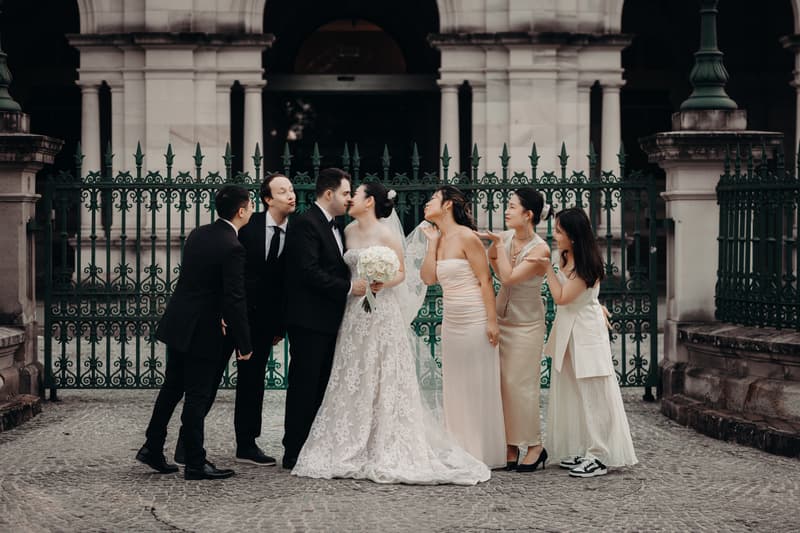 Aria and Antony stand in front of a green wrought iron fence at Queensland Parliament building, surrounded by three bridesmaids in light-colored dresses and two groomsmen in black suits, posing playfully with the bride and groom.