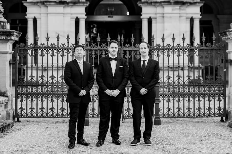 Antony, the groom, stands in the center with two men in suits in front of an ornate iron fence at Queensland Parliament building.