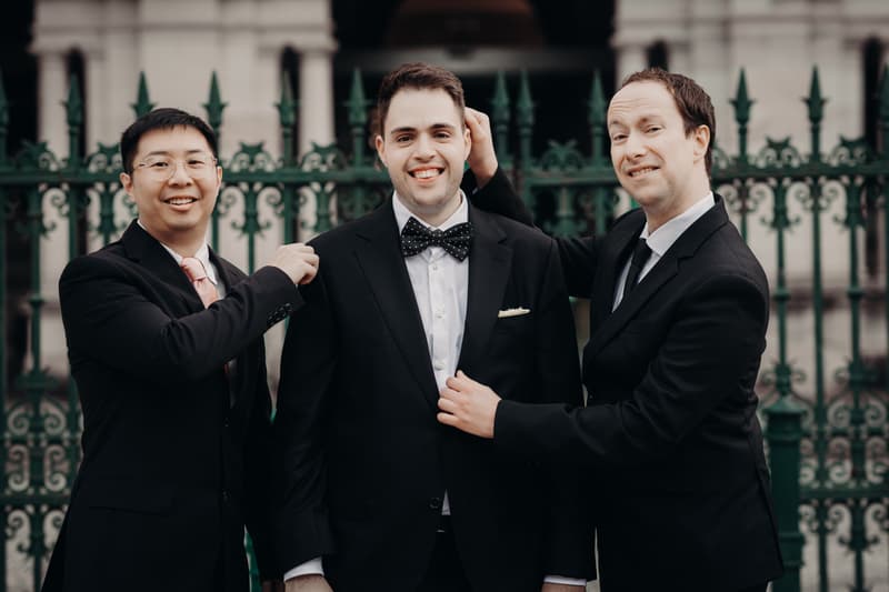 Antony, the groom, stands between two men in suits in front of a green iron fence at Queensland Parliament building. The man on the left adjusts Antony's jacket, while the man on the right playfully touches Antony's hair.