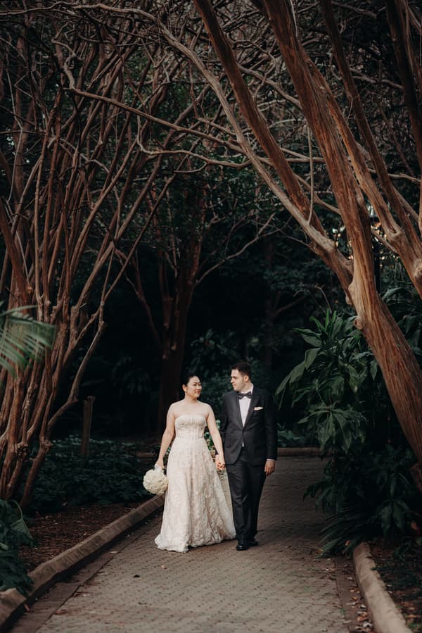 The bride Aria and the groom Antony walk hand in hand along a paved path surrounded by trees and greenery at City Botanic Gardens during their couple portraits session.