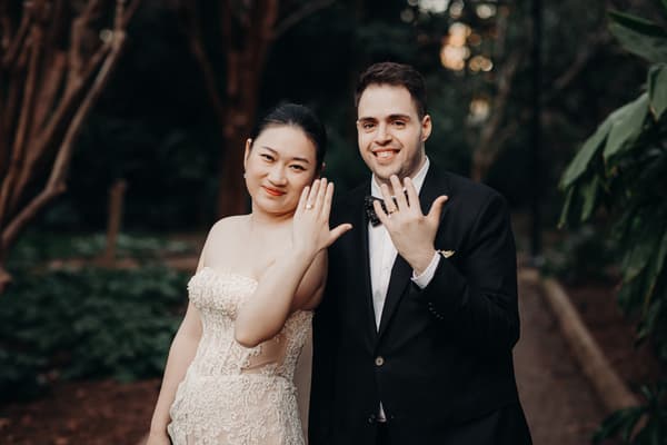 The bride Aria and the groom Antony stand together in the City Botanic Gardens showing their wedding rings to the camera.