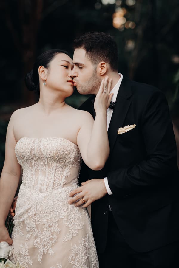 The bride Aria and groom Antony share a close moment, about to kiss, during their couple portraits session at City Botanic Gardens. Aria wears a strapless lace wedding gown and holds a bouquet, while Antony is dressed in a black suit with a bow tie and pocket square.